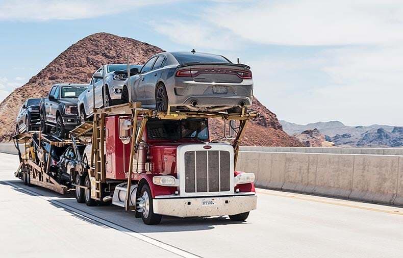 Loaded American car carrier hauling vehicles past a desert reservoir